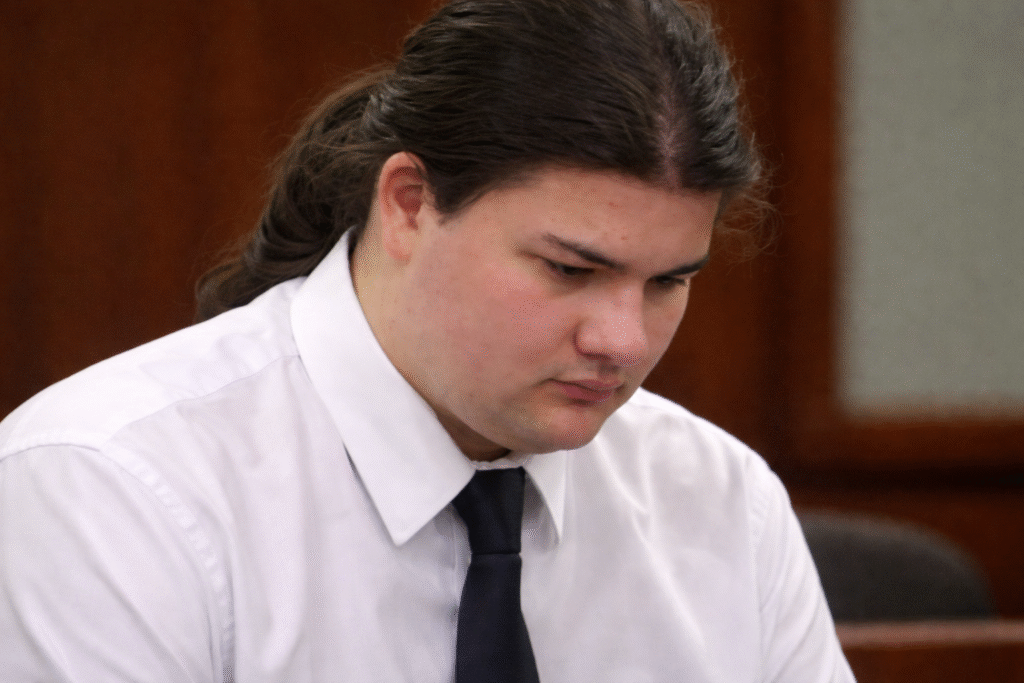 Close-up of a person in a white shirt and black tie seated in a courtroom, looking down with a serious expression against a blurred indoor background.