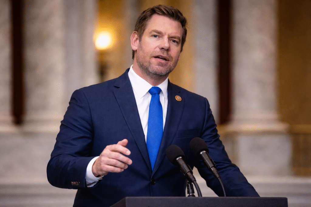 A man in a navy suit and blue tie speaks behind a podium with microphones, gesturing as he addresses an audience in a formal indoor setting.