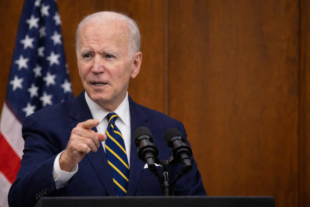 Joe Biden speaking at a podium with microphones, pointing forward, with a blurred American flag and wooden backdrop behind him