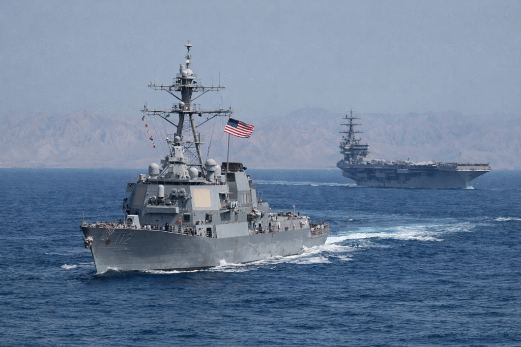 U.S. Navy destroyer sailing in open water with an aircraft carrier in the background near a mountainous coastline