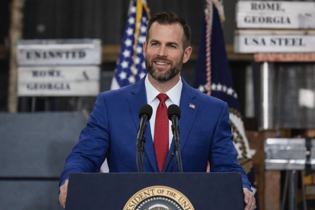 Bearded man in a blue suit speaking at a podium with microphones, American flags behind him, resembling a political speech or campaign event