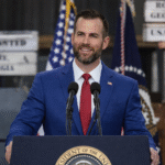 Bearded man in a blue suit speaking at a podium with microphones, American flags behind him, resembling a political speech or campaign event