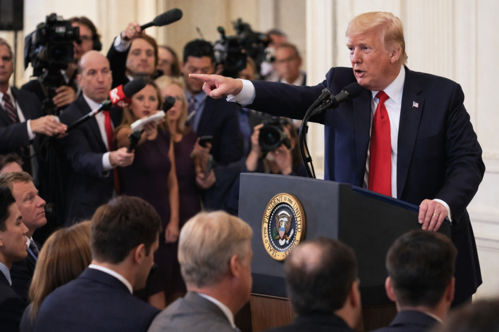 President Trump speaking at a podium pointing toward seated CNN staff, while reporters behind them extend microphones and cameras during a press conference