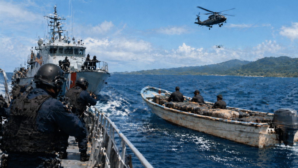 Coast guard vessels intercept a small unmarked boat carrying cargo on open ocean waters, with armed officers preparing to board and a helicopter overhead near a tropical coastline