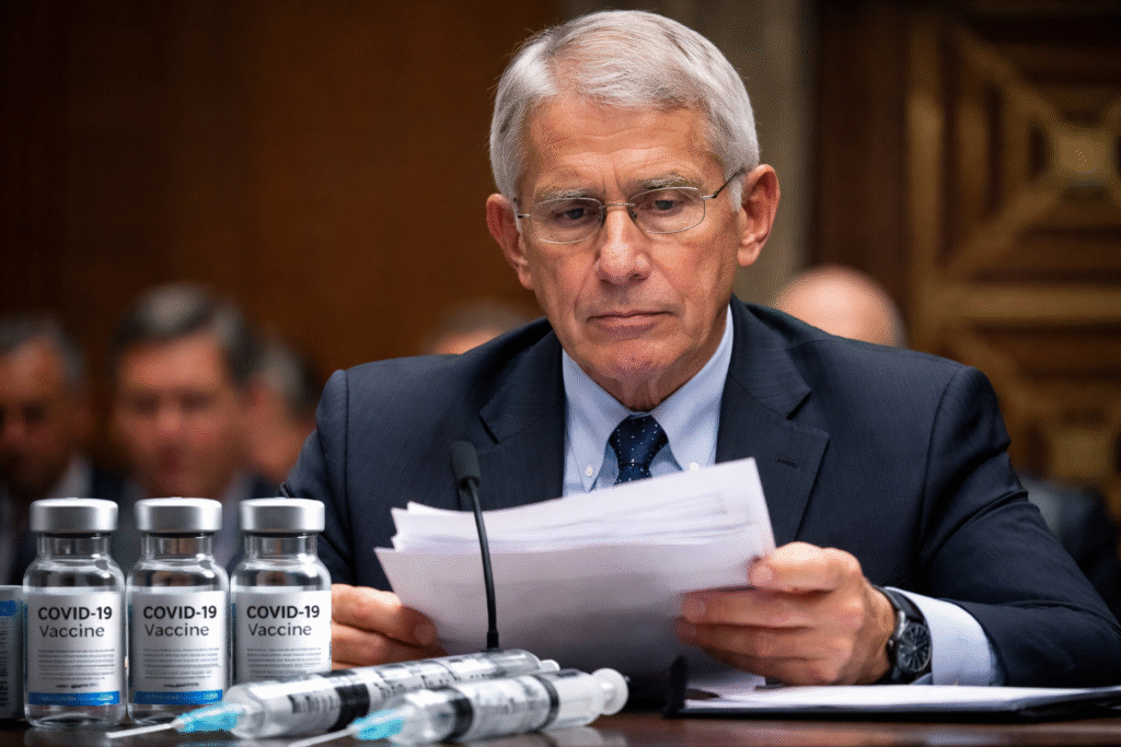 Dr. Anthony Fauci seated at a hearing table reviewing documents, with medical vials and syringes related to COVID-19 vaccines placed in front of him