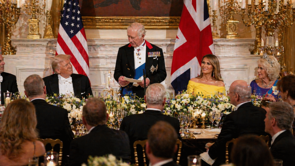 Formal state dinner with dignitaries seated at a decorated table, a speaker standing to give remarks, and American and British flags displayed behind them