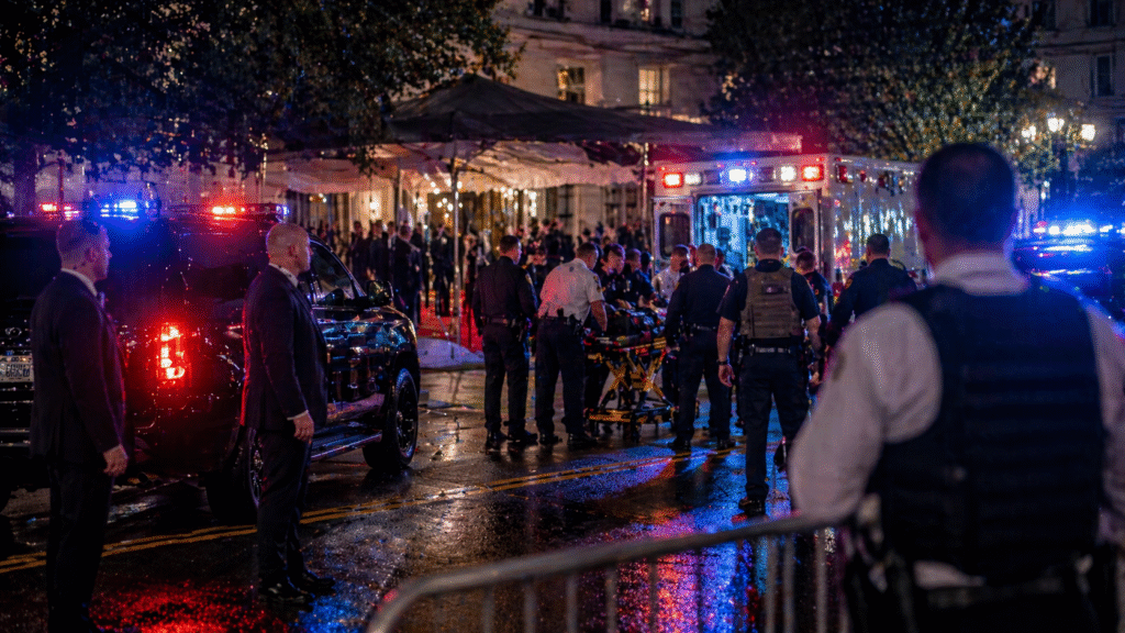 Emergency responders and law enforcement gather outside a large event venue at night, surrounding an ambulance with flashing lights as officers secure the area and assist an injured individual.