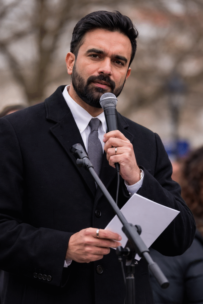 Man in a dark coat speaking into a microphone at an outdoor event, holding a paper with a serious expression while people stand blurred in the background.