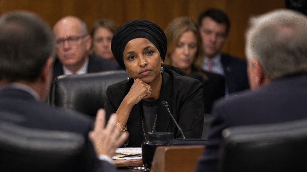 Female politician seated at a desk during a formal government hearing, listening intently as officials question her in a committee room