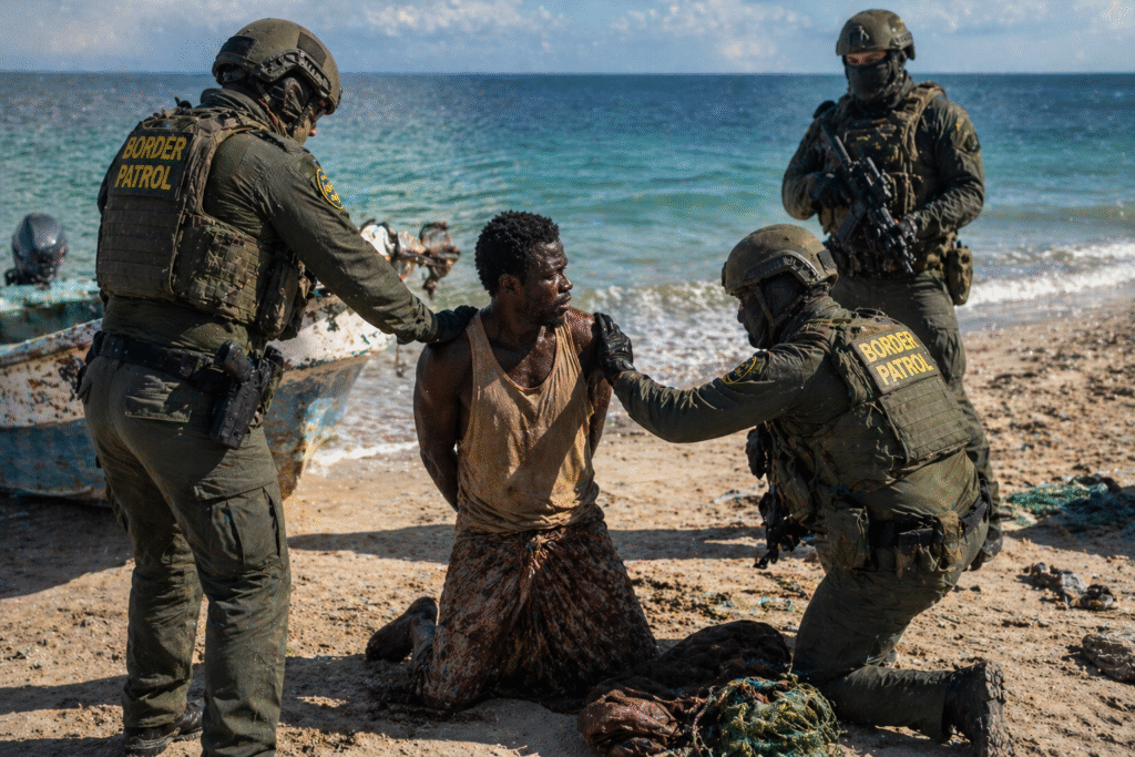 Man kneeling on a beach with hands restrained while armed officers secure him near the shoreline, with a small boat and ocean in the background
