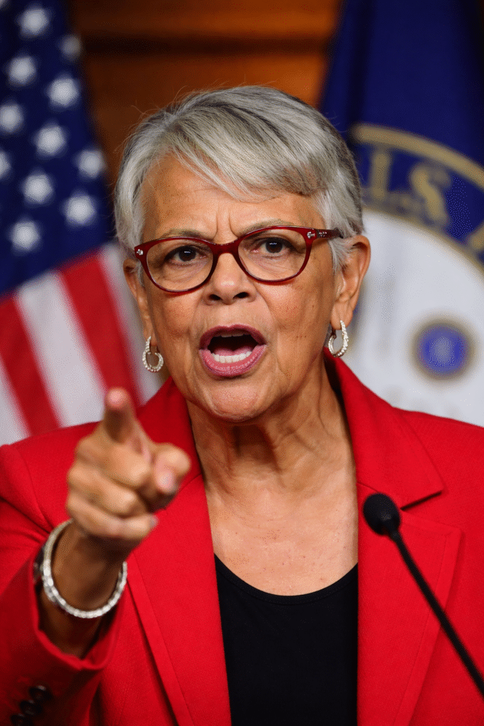 Close-up of a congresswoman in a red blazer speaking at a podium, pointing forward with an American flag blurred in the background.