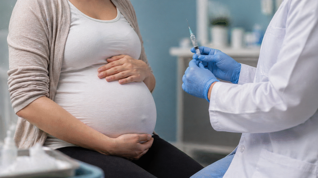 Pregnant woman sitting in a clinical exam room while a healthcare worker wearing gloves prepares a syringe for vaccination