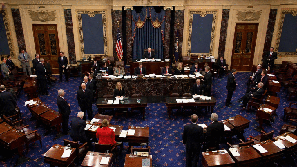 Interior of a legislative chamber with lawmakers gathered, desks arranged across a blue-carpeted floor, and a central podium beneath American flags during a formal session.