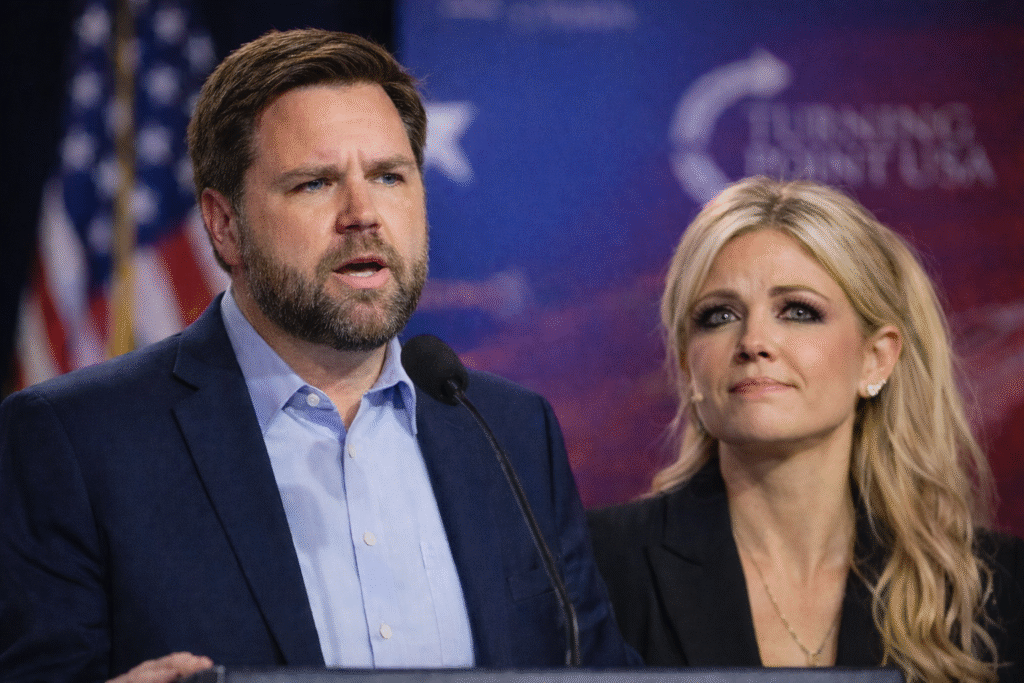 JD Vance speaking at a podium with a microphone, while a blonde woman stands beside him looking directly toward the camera, set against a blurred backdrop with an American flag.