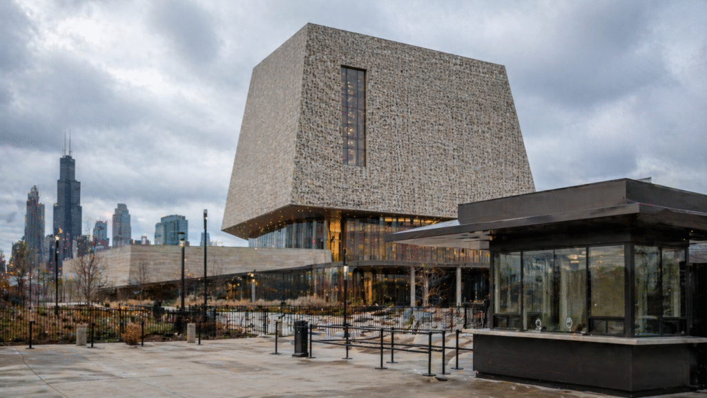 Modern museum-style building with a ticket booth in the foreground under an overcast sky, set against a city skyline resembling Chicago