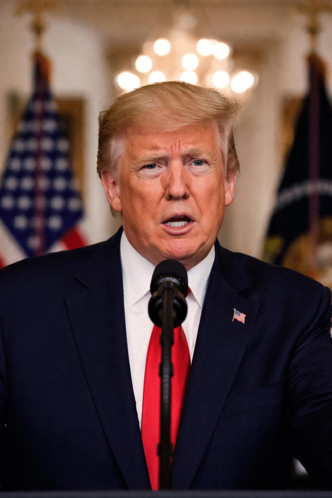 Close-up of President Donald Trump delivering a formal address at the White House podium, wearing a navy suit and red tie, with blurred American flags in the background
