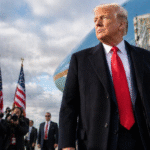 President Donald Trump standing on an airport tarmac near a presidential aircraft with American flags and photographers in the background