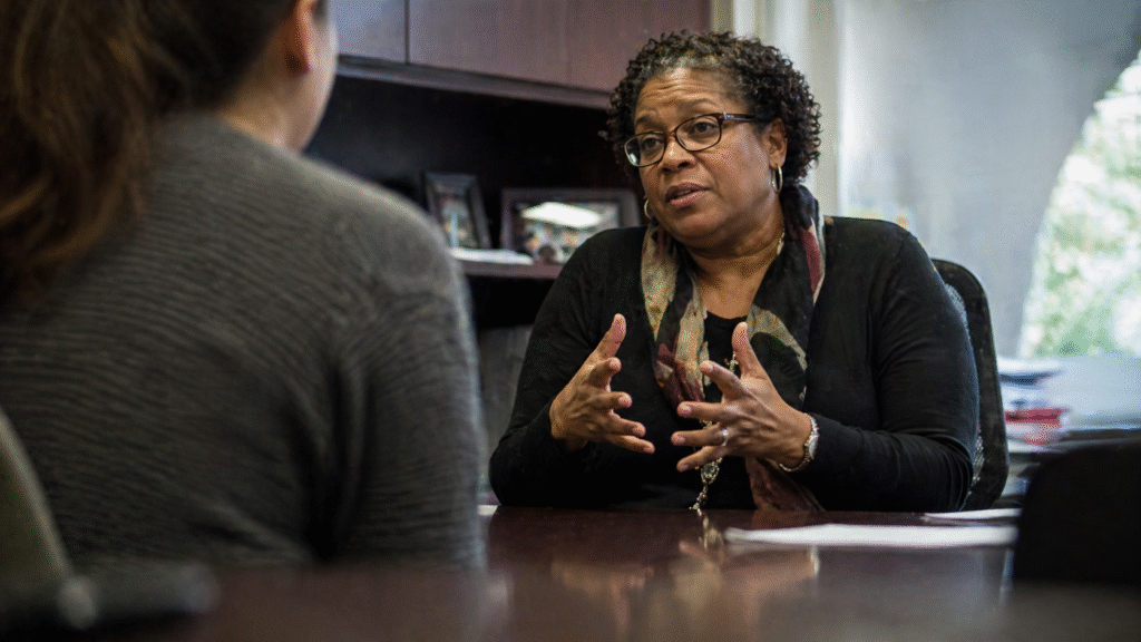 University administrator speaks across a desk in an office setting, gesturing while explaining a point, with another person seated opposite in a professional academic environment.