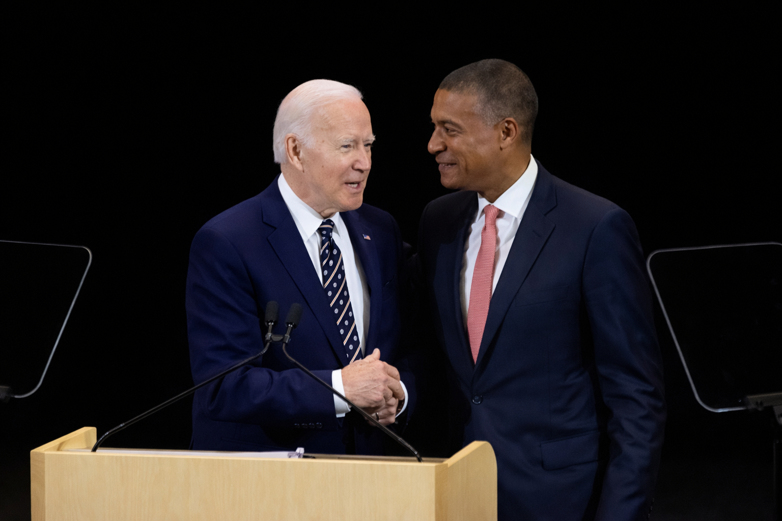 Two men in suits stand closely behind a podium, shaking hands and speaking quietly, with microphones visible and a dark, minimal background.