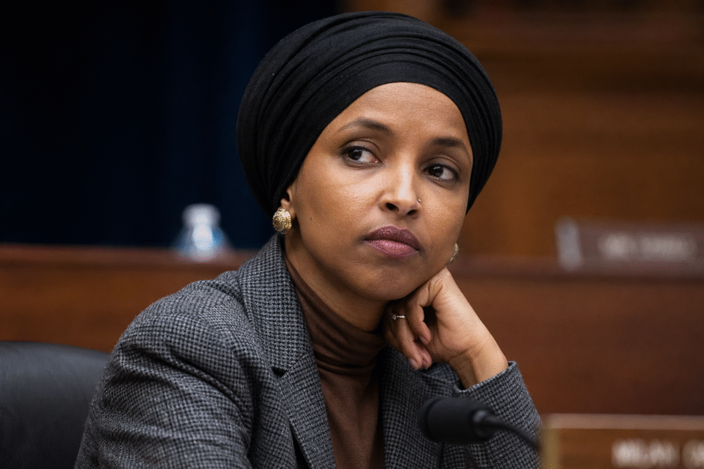 Close-up of a female politician seated in a formal hearing room, wearing professional attire and a head covering, with a thoughtful expression and blurred government setting in the background.