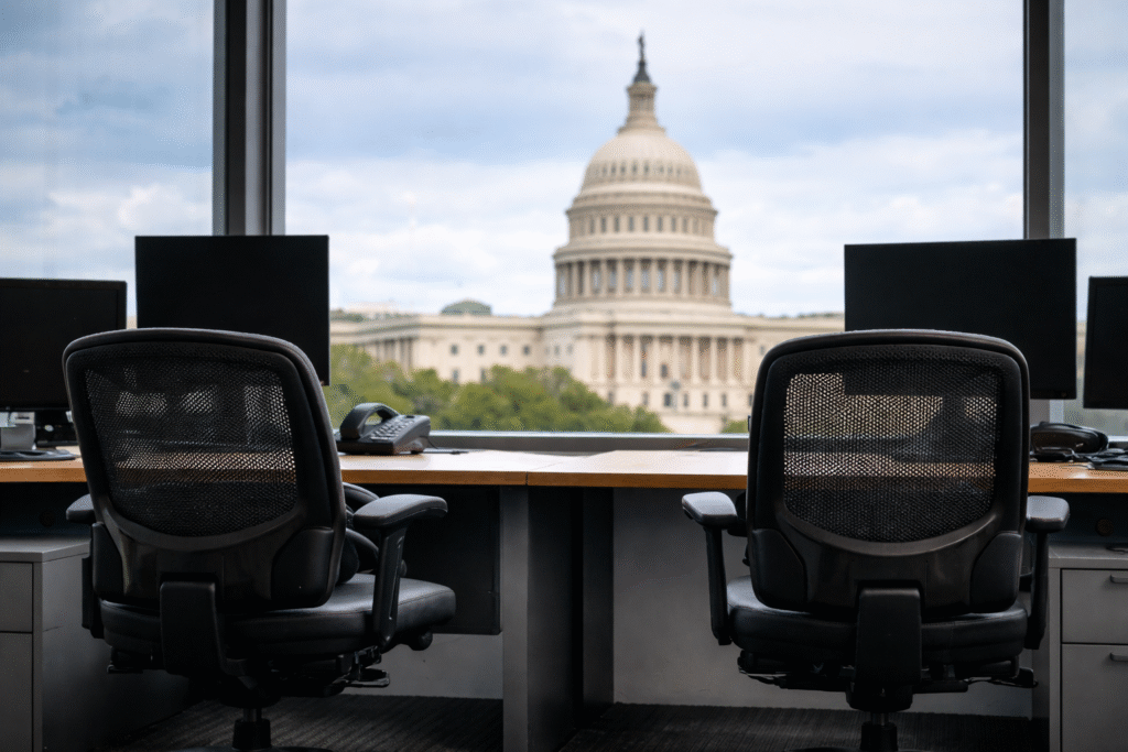 Empty office desks and chairs with the U.S. Capitol building visible in the background, symbolizing a reduced federal workforce.