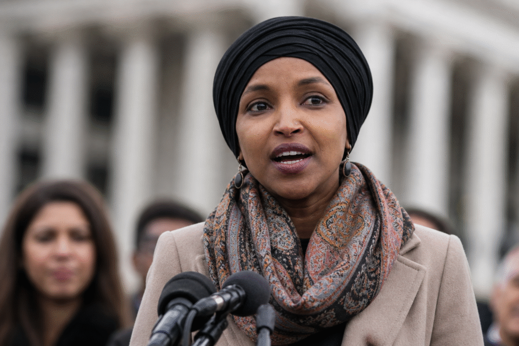 A woman wearing a dark headwrap and patterned scarf speaks at an outdoor podium with microphones, with blurred figures and a large building in the background.