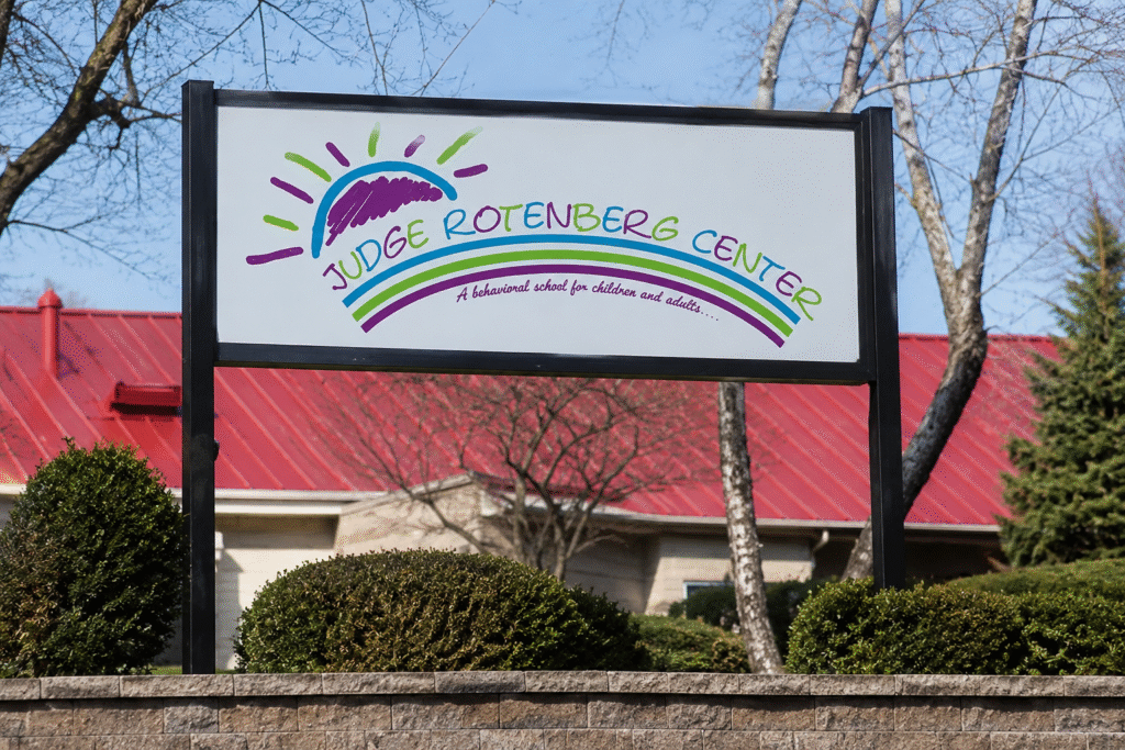 Sign outside the Judge Rotenberg Center behavioral facility in Massachusetts with a red-roofed building and trees in the background.