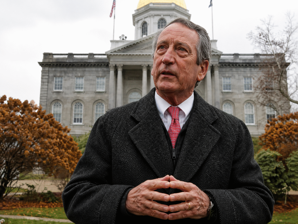 Older man in a dark coat standing outdoors in front of a neoclassical government building, hands clasped together, with autumn trees in the background